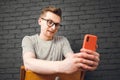 Young man in glasses looking at smartphone while sitting on a chair on a background of black brick wall. Copy, empty space for Royalty Free Stock Photo