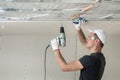 Young man in goggles fixing drywall suspended ceiling to metal frame using electrical screwdriver on ceiling insulated with shiny