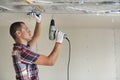 Young man in goggles fixing drywall suspended ceiling to metal frame using electrical screwdriver on ceiling insulated with shiny