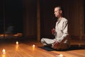 A young man in gray practice clothes sits in a lotus position with a red rosary in his hand in meditation. Dark room