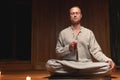 A young man in gray practice clothes sits in a lotus position with a red rosary in his hand in meditation. Dark room