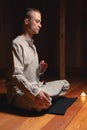 A young man in gray practice clothes sits in a lotus position with a red rosary in his hand in meditation. Dark room