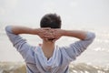 Young man with hands behind head, looking out to sea