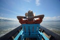 Young man in hat sitting on chair in boat with hands behind head and enjoying life, blue sky and clean water of lake Royalty Free Stock Photo