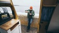 Young man drinking tea from thermos in front of camper van with a lake on background