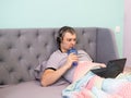 A young man in headphones lies reclining in a bed and watches video or works on a laptop. He holds a plastic glass with a tube and