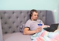 A young man in headphones lies reclining in a bed and watches video or works on a laptop. He holds a plastic glass with a tube and