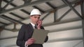 A young man in a helmet with documents speaks on a walkie-talkie at a construction site. The boss in a suit keeps