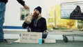 Young man helps to homeless person and giving him some food while beggar drink alcohol and sit near shopping cart at Royalty Free Stock Photo