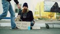 Young man helps to homeless person and giving him some money while beggar drink alcohol and sit near shopping cart at Royalty Free Stock Photo