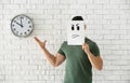 Young man hiding face behind sheet of paper with drawn emoticon against white brick wall with clock Royalty Free Stock Photo