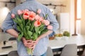 The young man is hiding flowers behind their backs to his girlfriend at home