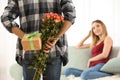 Young man hiding gift and flowers for girlfriend behind his back at home