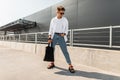 Young man hipster in stylish clothes in sunglasses with a black cloth bag in vintage sandals stands is in a city Royalty Free Stock Photo