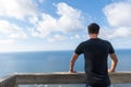 Young man with his back to a wooden gazebo, looking at a sunny seascape with white clouds, horizontal