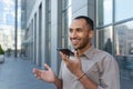 Young man hispanic standing outside, holding phone, talking through loudspeaker, smiling Royalty Free Stock Photo