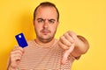 Young man holding blue credit card standing over isolated yellow background with angry face, negative sign showing dislike with Royalty Free Stock Photo