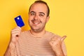 Young man holding blue credit card standing over isolated yellow background pointing and showing with thumb up to the side with Royalty Free Stock Photo
