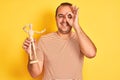 Young man holding figure of art dummy standing over isolated yellow background with happy face smiling doing ok sign with hand on Royalty Free Stock Photo