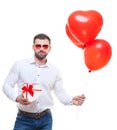 Young man holding gift box and balloons over white background. with glasses in the shape of heart. Looking at camera Royalty Free Stock Photo