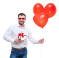 Young man holding gift box and balloons over white background. with glasses in the shape of heart. Looking at camera Royalty Free Stock Photo