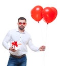 Young man holding gift box and balloons over white background. with glasses in the shape of heart. Looking at camera Royalty Free Stock Photo