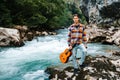 Young man holding guitar standing on the bank of a mountain river on a background of rocks and forest. Handsome hippie style