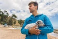 Young man holding his adopted mixed Dalmatian puppy in scenic place with beach, sky and sea on background. Pets adoption concept Royalty Free Stock Photo