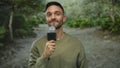 Young man holding microphone during interview in lush park setting, surrounded by greenery, showcasing casual attire and engaging Royalty Free Stock Photo