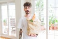 Young man holding paper bag of fresh groceries from the supermarket scared in shock with a surprise face, afraid and excited with Royalty Free Stock Photo