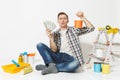Young man holds bundle of dollars, cash money, sits on floor with paint can, instruments for renovation apartment Royalty Free Stock Photo
