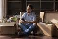 Young man holds smartphone sits on floor near cardboard boxes