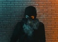 Young man in hoodie and balaclava in front of brick wall looking down and blow smoke out