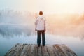 Young man in hoodie, hat and pants standing on wooden pier on pond shore with melancholy fog at sunrise. Czech morning landscape Royalty Free Stock Photo