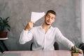 A young man in a hot office fanns himself with a piece of paper. A guy in a shirt sitting at a table, a tired worker is Royalty Free Stock Photo
