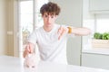 Young man investing money using piggy bank at home with angry face, negative sign showing dislike with thumbs down, rejection Royalty Free Stock Photo