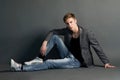 Young man in jacket and jeans sitting on dark background