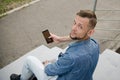 Young man in jeans and a denim shirt is sitting on the stairs with a smartphone in his hand turned around and looks directly at