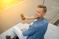 Young man in jeans and a denim shirt is sitting on the stairs with a smartphone in his hand turned around with smiling and looks