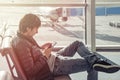 Young man in jeans and jacket sits on chair spend time by using mobile phone in airport lounge. Booking hotel in foreign country. Royalty Free Stock Photo