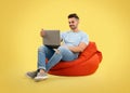 Young man with laptop on bean bag chair, yellow background Royalty Free Stock Photo