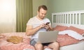 Young man with a laptop in bed working at home. Cat siiting near him.