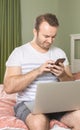 Young man with a laptop in bed working at home. Cheerful young adult with phone in hands