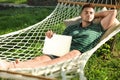 Young man with laptop resting in comfortable hammock at garden Royalty Free Stock Photo