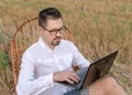 Young man with laptop sitting in a rocking chair outdoors. Handsome caucasian man with glasses working remotely outdoors