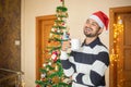 Young man laughing enjoying coffee wearing santa hat celebrating christmas alone at home with decorated tree and lights in Royalty Free Stock Photo