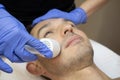 Young man laying on a facial therapy couch covered with the white towel with opened eyes. Facial therapist hands holding