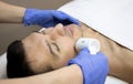 Young man laying on a facial therapy couch covered with the white towel with opened eyes. Facial therapist hands holding