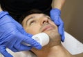 Young man laying on a facial therapy couch covered with the white towel with opened eyes. Facial therapist hands holding