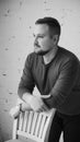 A young man leaning on the back of a chair looks to the side against a white brick wall. Black and white photo